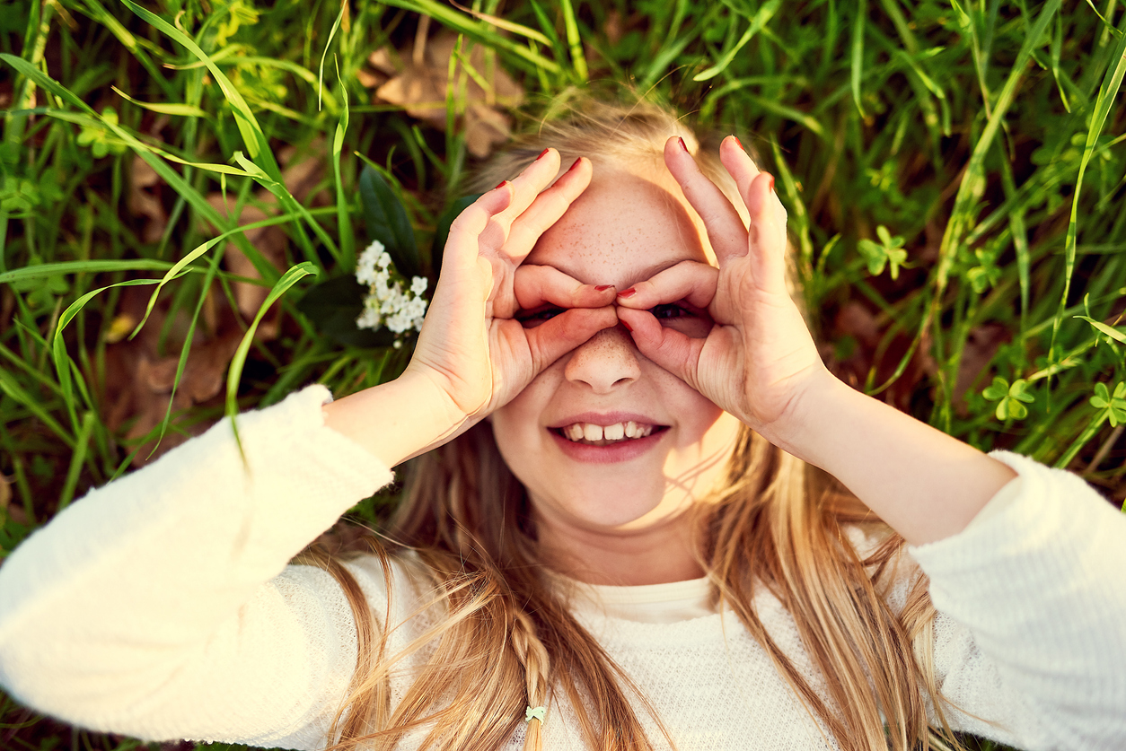 Smiling child lying in grass and looking through finger binoculars, enjoying outdoor play without eye allergy symptoms